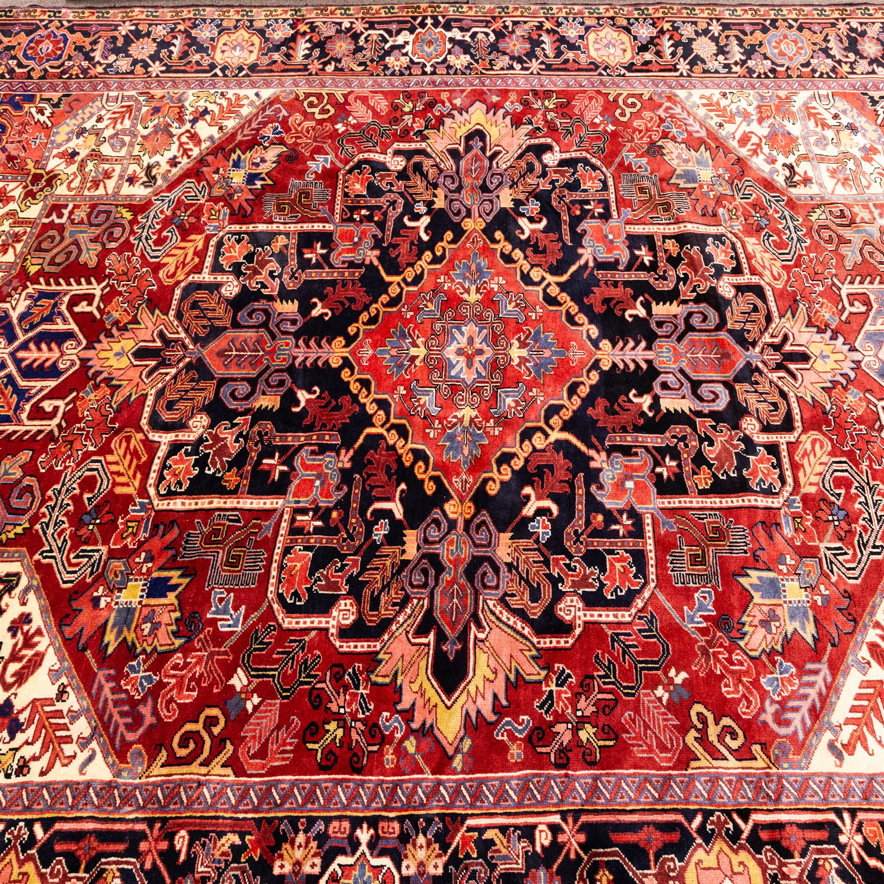 North West Persian Heriz hand-knotted carpet, madder red field with large central stepped medallion with angular floral and geometric designs, surrounded by stylized palmettes, leaves and tribal motifs with indigo corner spandrels, enclosed by a broad navy border of geometric floral devices between multiple guard stripes, with cotton fringe ends 