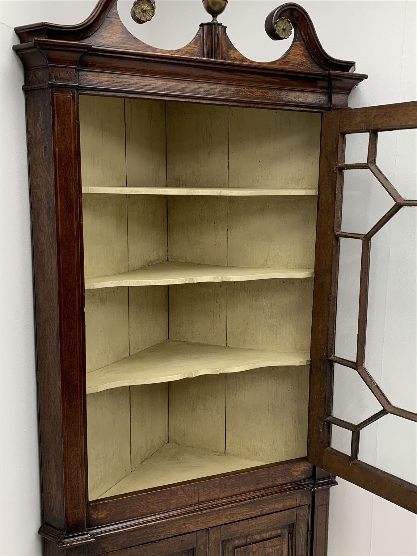 George III oak floor standing corner cabinet, scrolled swan neck pediment with central finial over astragal glazed door, painted interior fitted with three shaped shelves, the lower section fitted with single shaped shelf enclosed by two fielded panel doors inlaid lozenges, on bracket feet, H195cm, W77cm