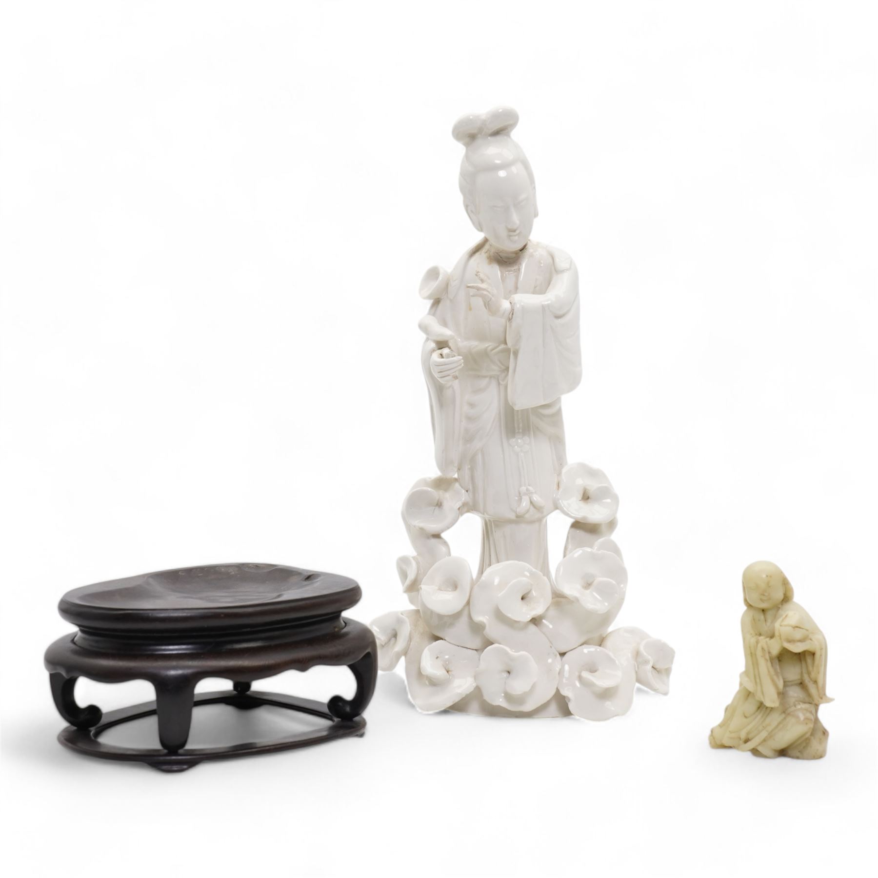 Chinese blanc de Chine figure of Guanyin, wearing robes and standing upon a lotus base, H25.5cm, a jichimu carved wooden stand, and carved greenstone figure of an immortal (3)
