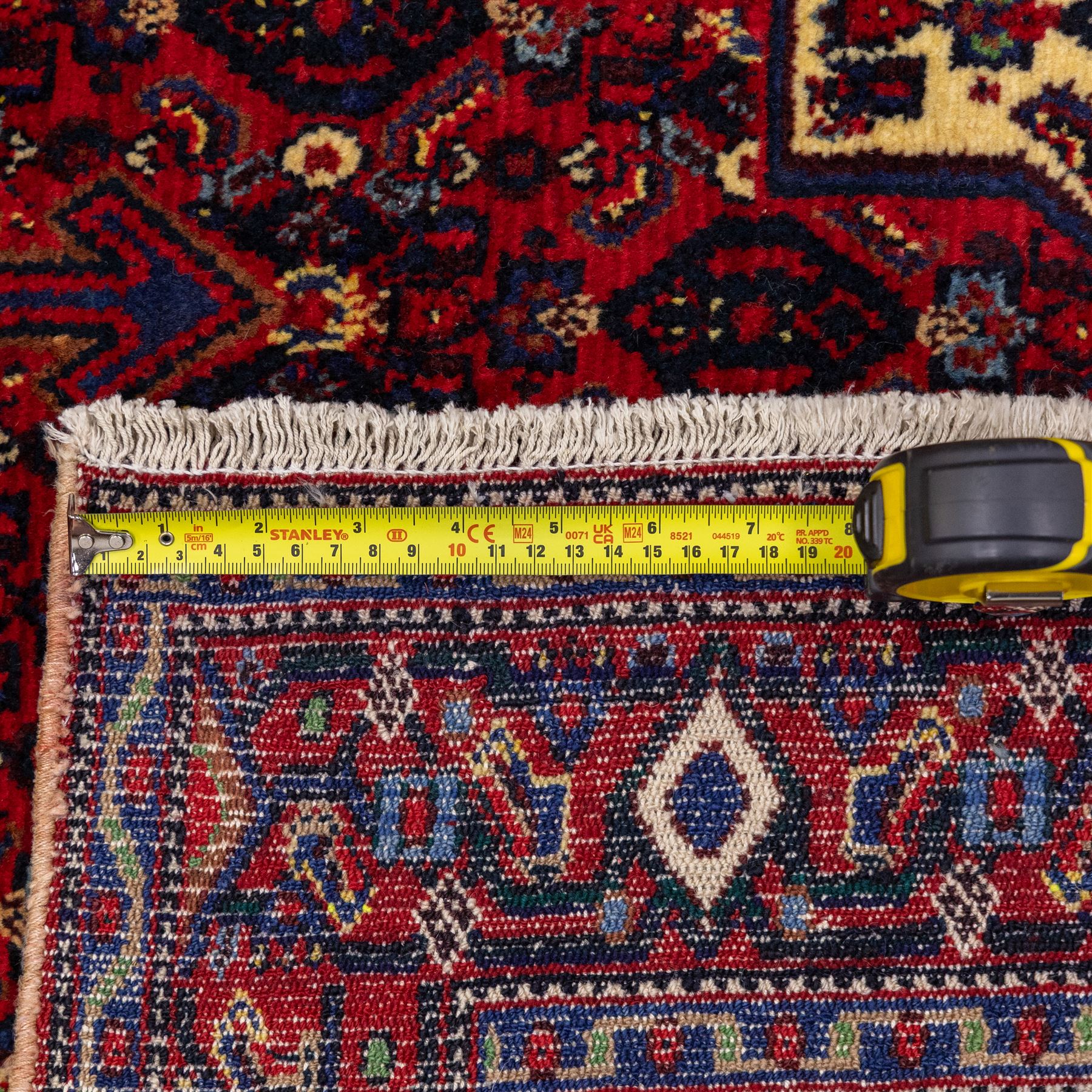 North West Persian Senneh hand-knotted rug, dark indigo field with a central bold stepped lozenge medallion of madder red and deep blue, enclosed within an ivory serrated contour forming a cruciform outline, medallion with repeating boteh and floral devices, field decorated with an all-over Herati pattern of rosettes enclosed by angular leaves principal border of madder red ground decorated with alternating floral heads, flanked by narrow guard stripes, with cotton fringe ends 