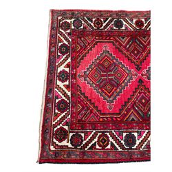 Persian Hamadan red ground rug, decorated with six stepped lozenge medallions, the guarded border with repeating stylised flower head decoration, with inscription to corner