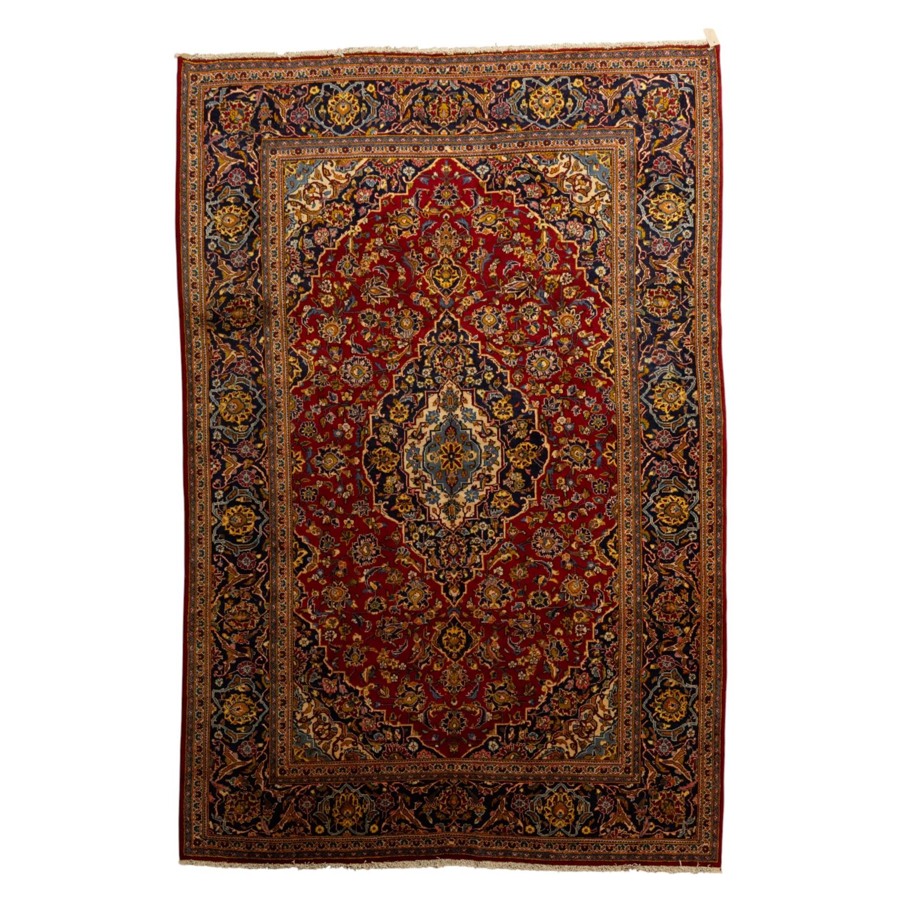 Central Persian Kashan hand-knotted carpet, crimson red field with a central shaped palmette medallion with pendants, filled with dense scrolling and floral motifs, enclosed by a navy blue border decorated with palmettes and meandering vine scrolls, flanked by multiple guard stripes, with cotton fringe ends 
