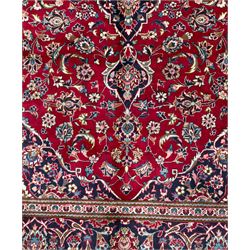 Central Persian Kashan hand-knotted carpet, the deep red ground with a central lozenge medallion in ivory and navy, surrounded by dense scrolling floral and vine motifs, navy spandrels and a wide border with stylised palmettes and flowerheads enclosed by multiple guard stripes