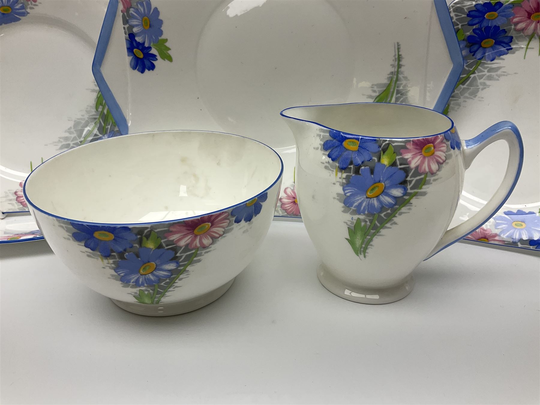 Shelley Strand shaped tea wares, decorated with blue and pink daisies upon a white ground, comprising four teacups, four saucers, three tea plates, milk jug, sugar bowl and cake plate, all with printed green mark beneath and painted pattern no 12216