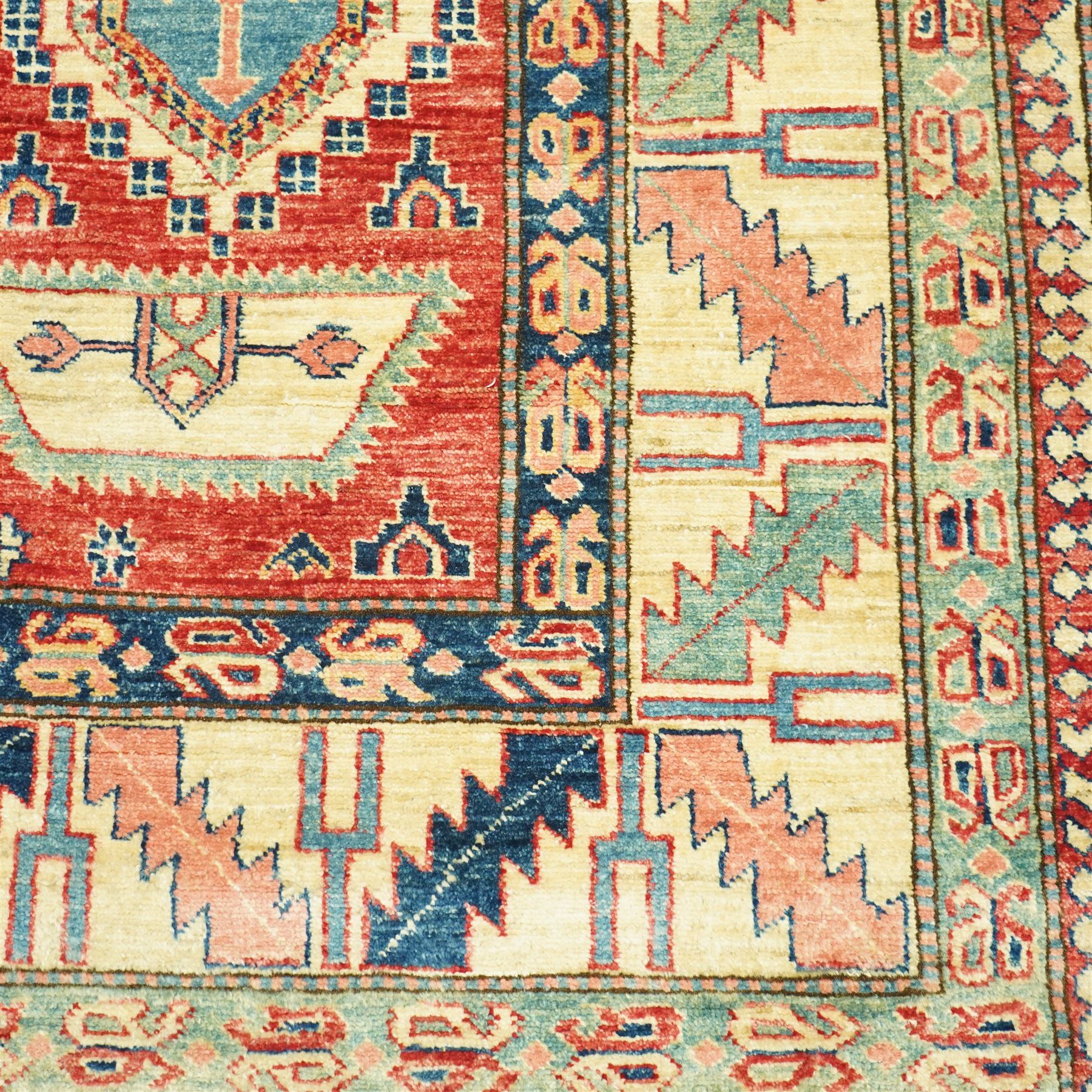 Caucasian Kazak hand-knotted rug, madder red field with bold geometric medallions and angular tribal motifs, within multiple patterned borders in a palette of indigo, red, ivory and green 