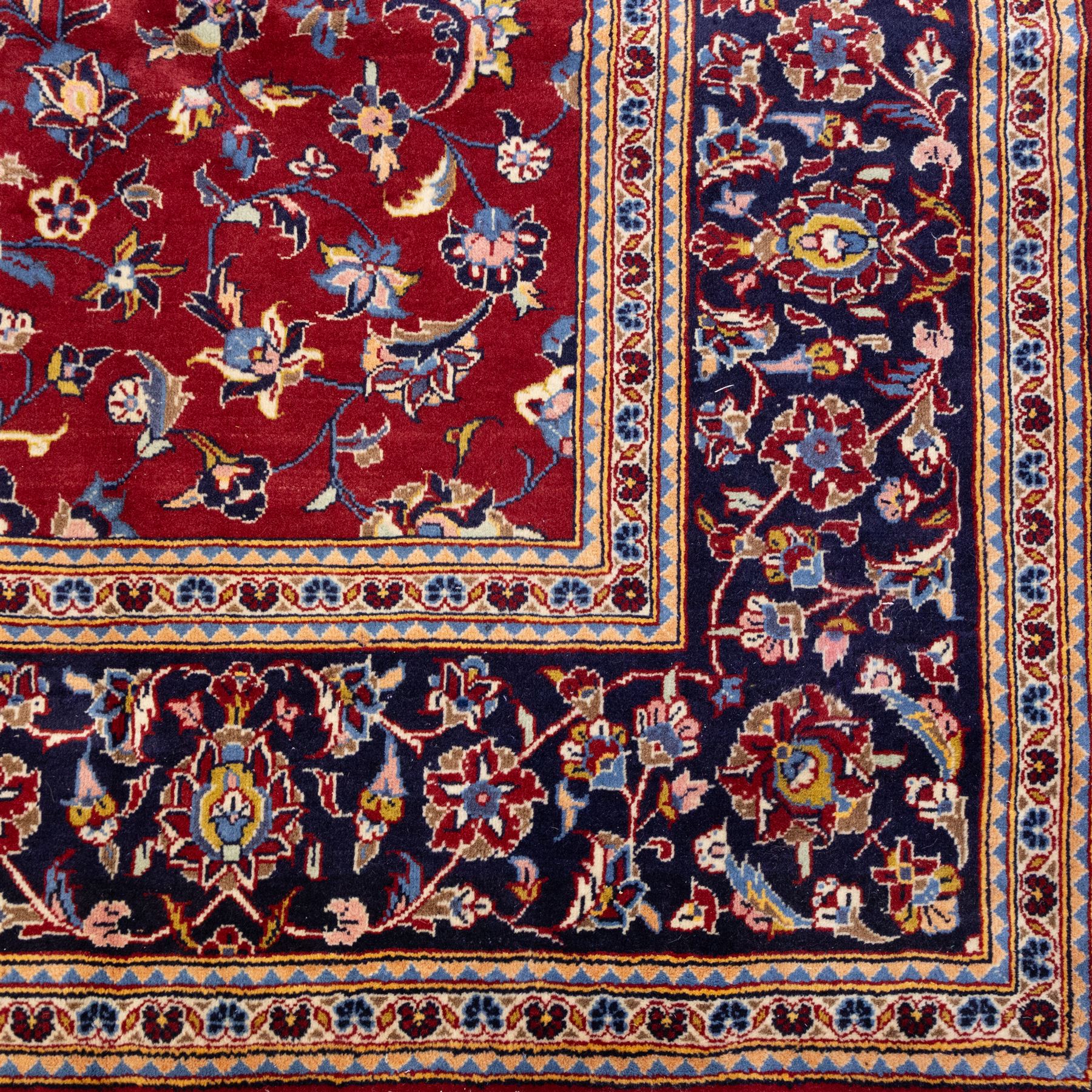 Central Persian Kashan hand-knotted rug, crimson field, decorated with an all-over floral and foliate design incorporating stylized palmettes, rosettes, and scrolling vine work, enclosed by a dark navy blue main border ornamented with flowering branches and palmettes flanked by multiple narrow guard borders with repeating floral motifs, with cotton fringe ends 