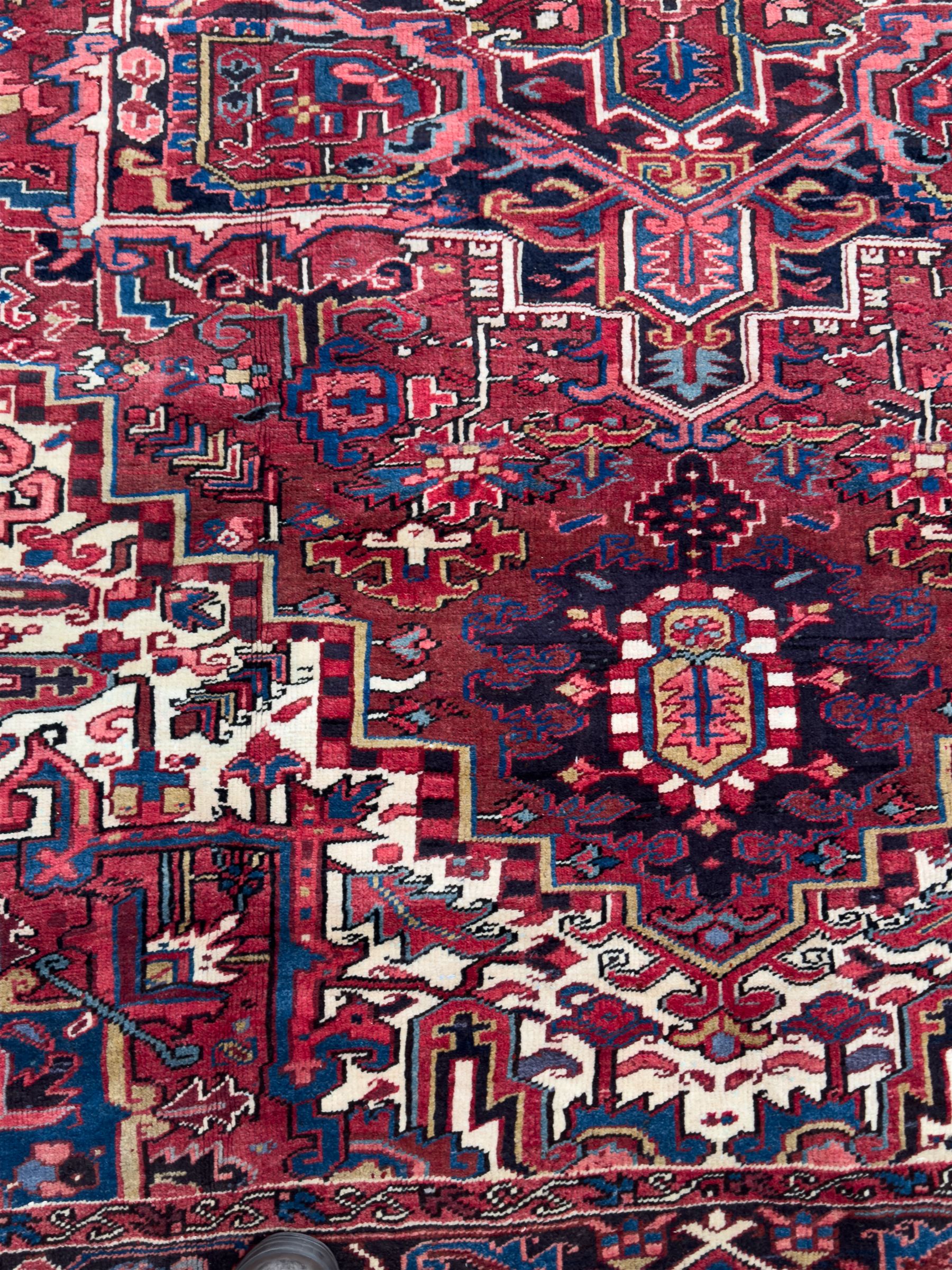 Northwestern Persian Heriz hand-knotted carpet, red madder field with a bold stepped medallion in indigo, ivory, and terracotta, flanked by angular floral motifs and stylized leaves, the spandrels set within a dark indigo border of rosettes and angular vine motifs between narrow guard stripes, 340cm x 245cm 