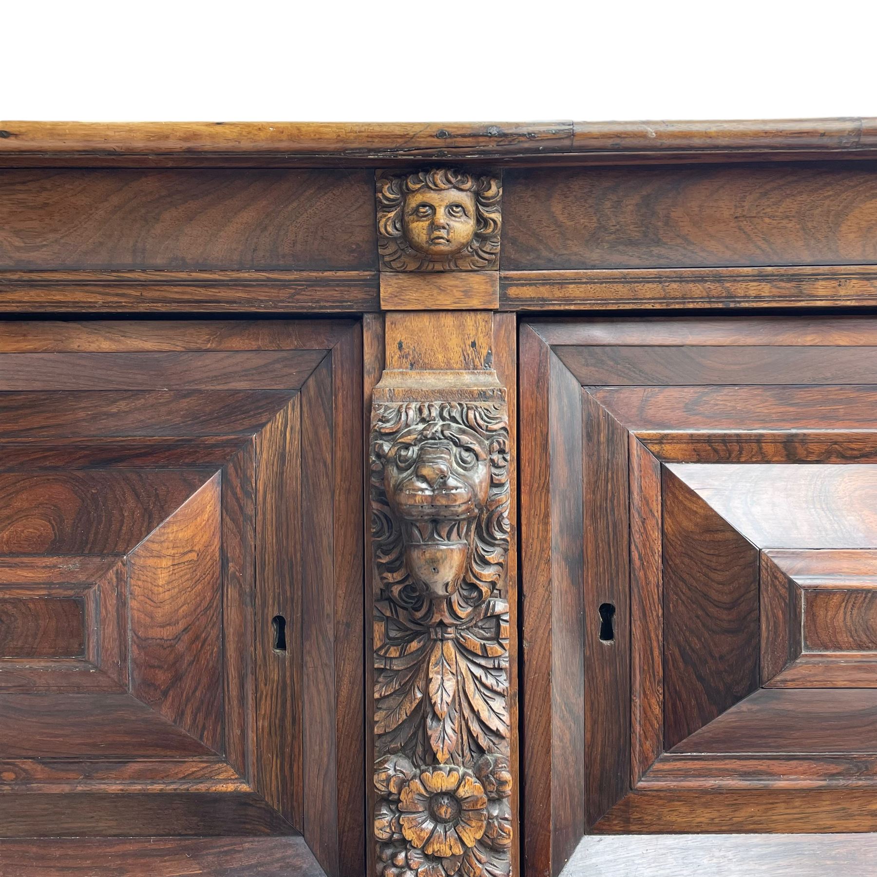 Late 17th century Flemish/Dutch rosewood and oak ‘Zeeuwse Kast’ or cupboard, projecting cornice carved with three putti masks, the uprights carved with lion masks over ribbons, flower heads and foliage, enclosed by four geometric panelled doors, lower central upright carved with flower head over foliate ribbon with bird among foliage, the escutcheon hidden by swivel berry carved cover, interior fitted with shelves, two drawers and shallow hinged cupboard, on two large turned and ebonised feet, circa 1680