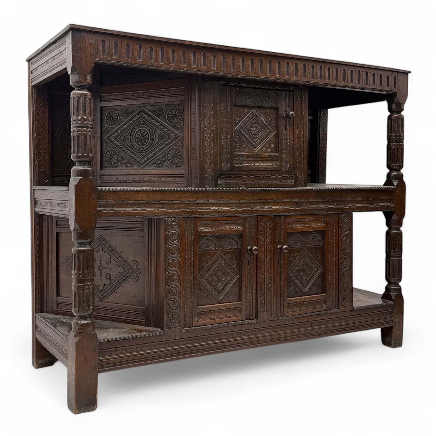 Early 20th-century  oak Jacobean two-tier revival court cupboard, rectangular top with carved notch and stylized foliate carved edge above a carved lozenge pattern frieze and a central cupboard with carved geometric designs and geometric carved panelled sides, above a carved panelled cupboard with two doors of similar geometric design, the whole flanked by two sets of stop-fluted baluster turned columns and supported on stile feet
