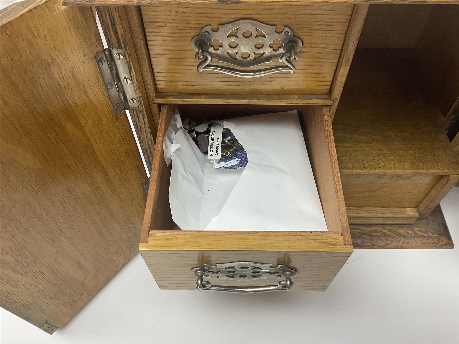Early 20th century oak smokers cabinet, with brass shield shaped mounts, opening to reveal a compartmentalised interior with three drawers, pipe rack and tobacco jar, together with a small wooden shelf, smokers cabinet H28.5cm