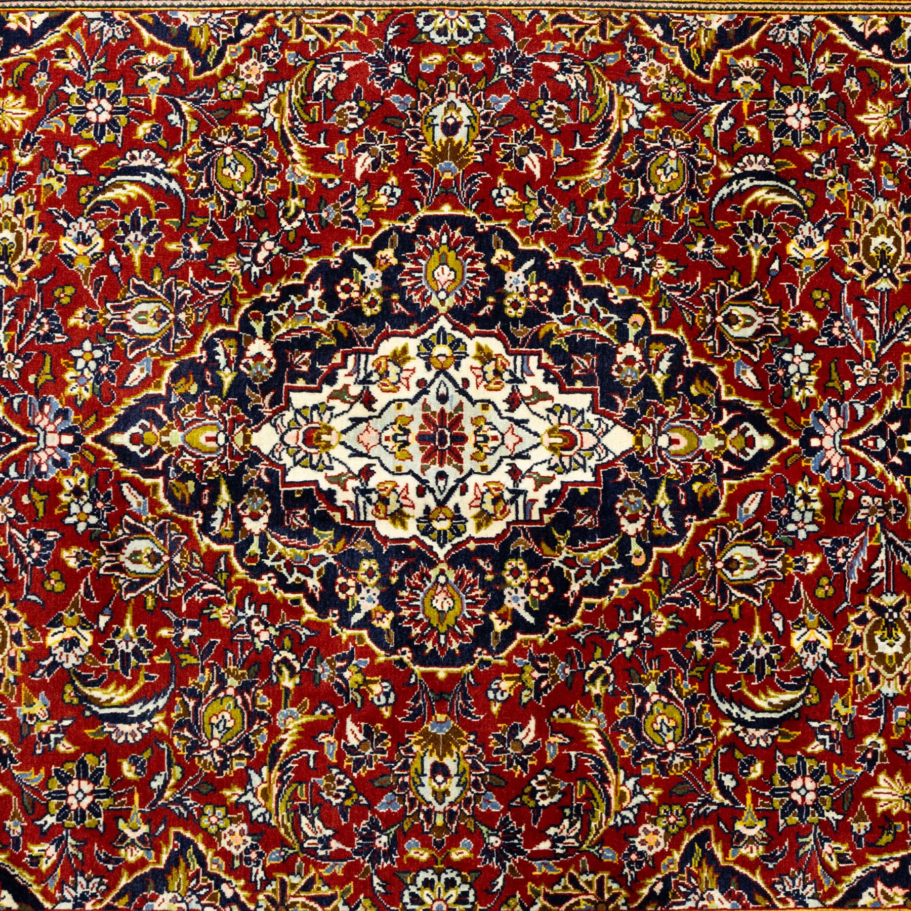 Central Persian Kashan hand-knotted carpet, crimson ground, with a central ivory shaped floral medallion with pendants, surrounded by dense scrolling foliate and floral motifs incorporating rosettes, palmettes and delicate vine work, enclosed by ivory spandrels and a dark navy border with repeating palmettes and meandering vines, flanked by floral guard stripes, with cotton fringes