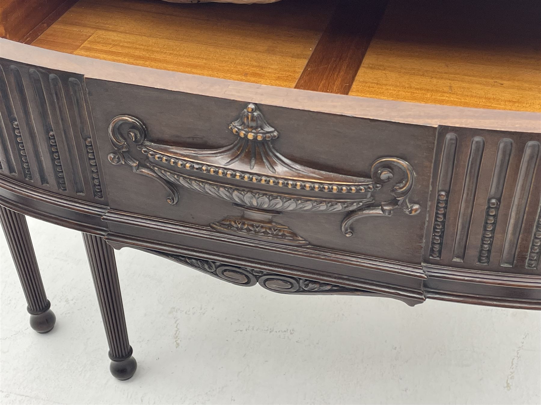 Early 20th century Hepplewhite style serving table, raised fleur-de-lis feather back with fluted detail and flower head mounts, the shaped top with carved foliate edge over single central drawer with applied urn motif, turned and reeded acanthus supports with recessed castors
