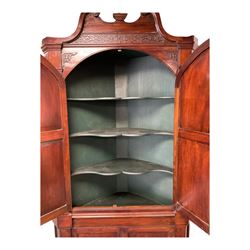 George III mahogany corner cabinet, the scrolled pediment surmounted by a brass finial and carved fretwork frieze, with fluted uprights flanking arched double doors over a painted interior fitted with shaped shelves, above a lower cupboard with twin panelled doors, on plinth base