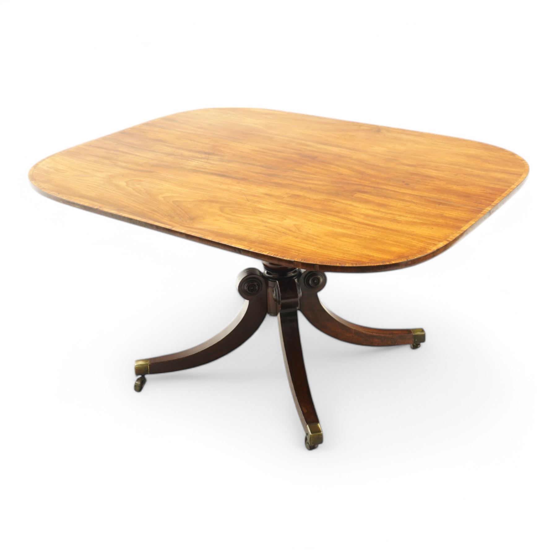 Regency figured mahogany breakfast table, figured rectangular tilt-top with rounded corners and rosewood banding, on turned column with four out-splayed supports with brass cups and castors 