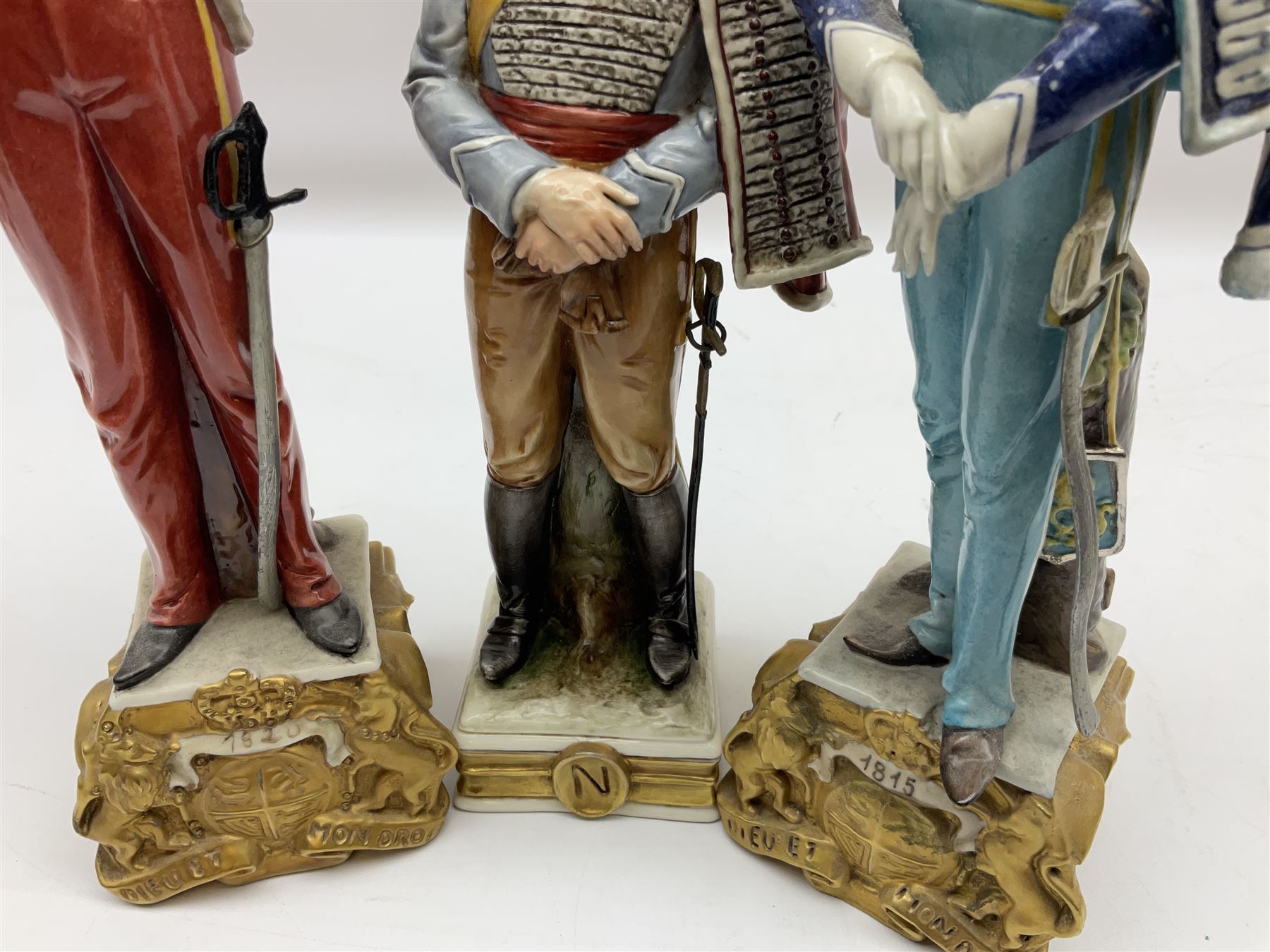 Group of seven Capodimonte figures of soldiers, to include four Bruno-Merli examples including Officer of the 12th Lancers 1820 and Captain of the 18th Hussars 1815, both raised upon ornate gilt plinths bearing British monarch motto in Latin 'Dieu et mon droit', and three further similar, all with crowned N mark beneath, tallest H32cm