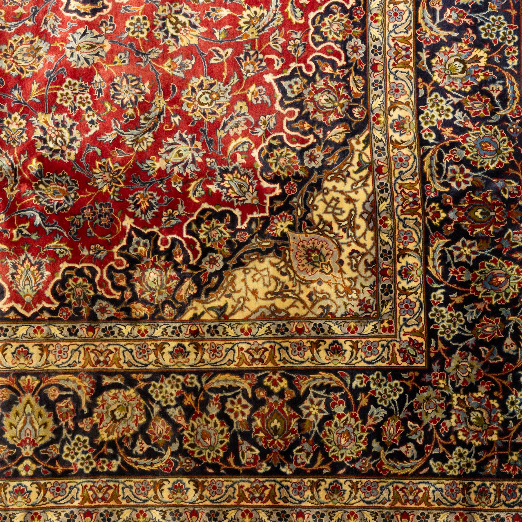 Central Persian Kashan hand-knotted carpet, crimson ground, with a large central ivory and dark navy floral medallion with pendants, surrounded by dense scrolling foliate and floral motifs incorporating rosettes, palmettes and vine work, enclosed by ivory spandrels and a dark navy border with repeating palmettes and meandering vines, flanked by floral guard stripes