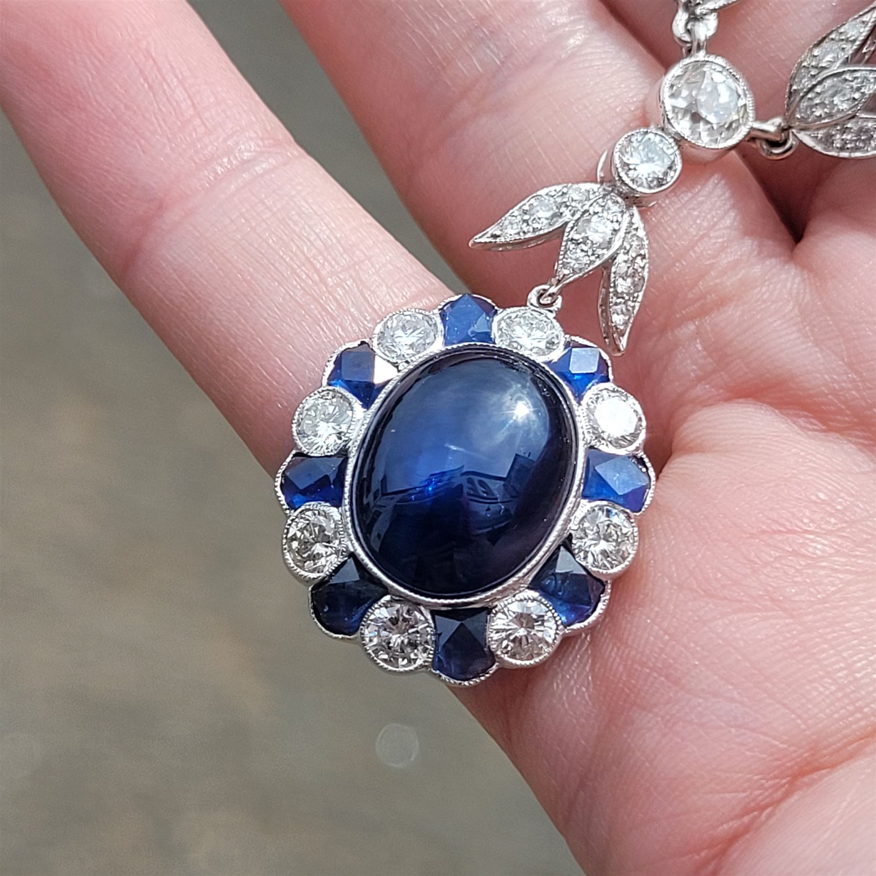 18ct white gold sapphire and diamond milgrain set necklace, oval cabochon cut sapphire surrounded by eight round brilliant cut diamonds and eight French cut sapphires, suspending from a diamond set leaf pendant, single round brilliant cut diamond and a larger old cut diamond, the chain set with six graduating cabochon cut sapphires, each surrounded by diamonds and spaced by a single diamond and a diamond set leaf, to a belcher link chain necklace, hallmarked, total sapphire weight approx 27.00 carat, total diamond weight approx 7.50 carat