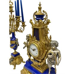 A 20th century continental gilt metal Lyre mantle clock with a pair of matching six light candelabra, spring driven movement housed in a gilt drum case on a brass and cobalt blue base with paw feet, two bronzed metal figures of mythological cherubs with animal legs and cloven hooves supporting a festooned garland swag, eight-day twin barrel striking movement with a floating lever balance escapement, striking the hours and half-hours on two bells, white enamel dial with roman numerals, minute track and pierced steel hands, dial inscribed “Imperial”. 