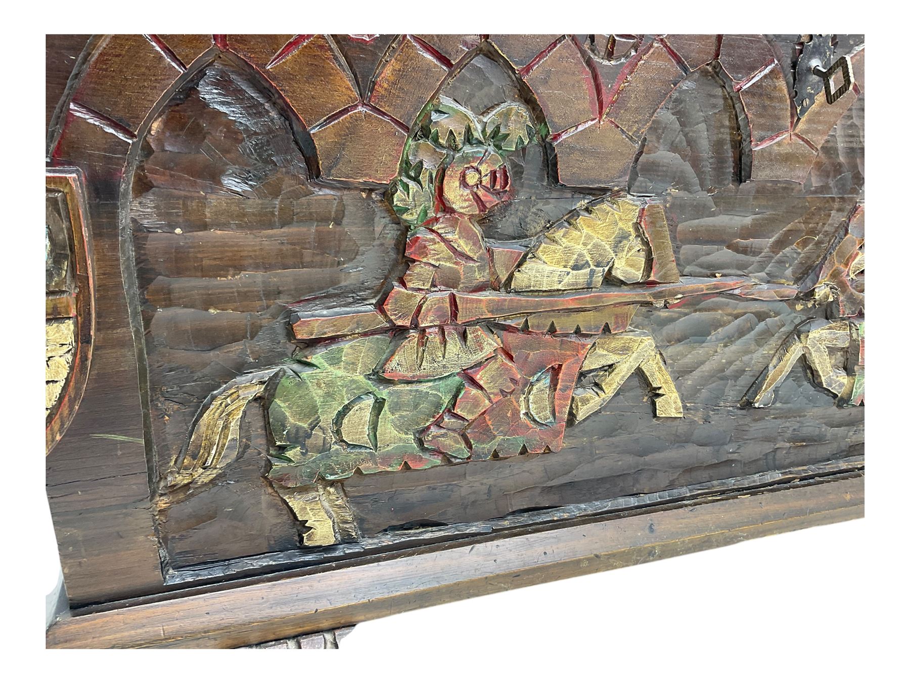Mid-20th century carved hardwood chest, the rectangular hinged top with decorative strap hinges, the front and sides deeply carved with medieval-style jousting knights, heraldic shields, and architectural elements, raised on shaped block feet