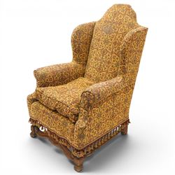 Pair of William & Mary design wingback armchairs, the arched high-back over rolled arms with a loose seat cushion and sprung seat, upholstered in old-gold ground foliate patterned fabric, raise on scroll feet united by a front serpentine stretcher