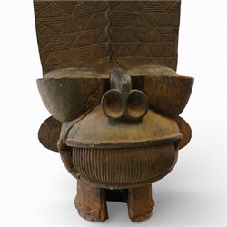 African carved hardwood ceremonial seat, monoxylous construction with twin dished seat wells, the tall back incised with repeating geometric triangular motifs, the front carved in the form of a tribal mask with projecting features, raised on a cylindrical base with ribbed and textured carving
