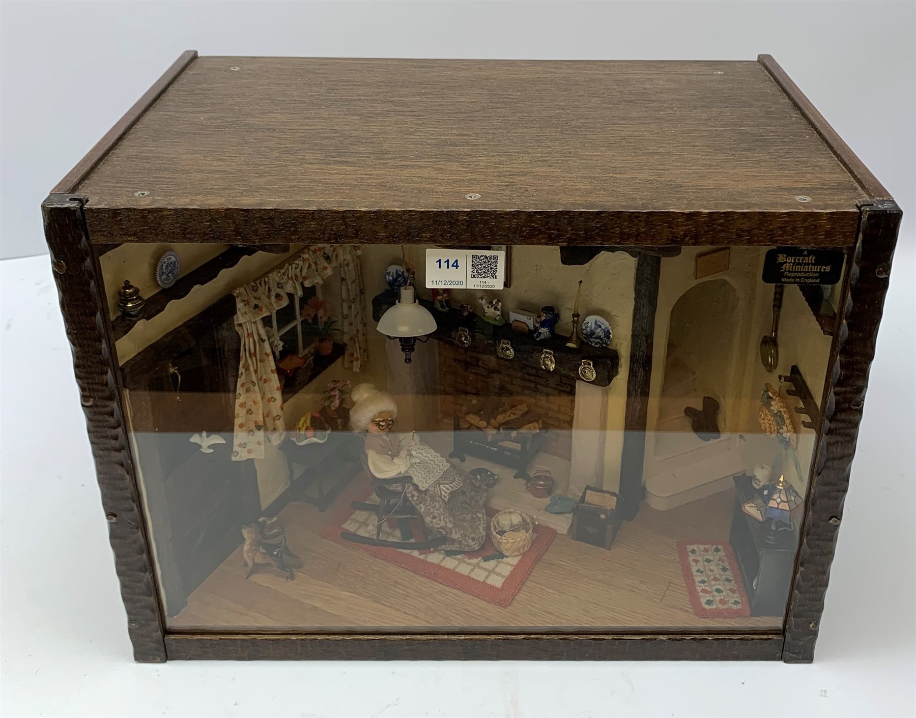 A 1/12th scale diorama of a cottage sitting room, depicting an elderly lady seated before a fireplace knitting, with dog and cat, in an interior furnished with blue and white plates, Staffordshire figures, toby jug, brass horse shoes, copper kettle, etc., H29cm L40cm D26cm.