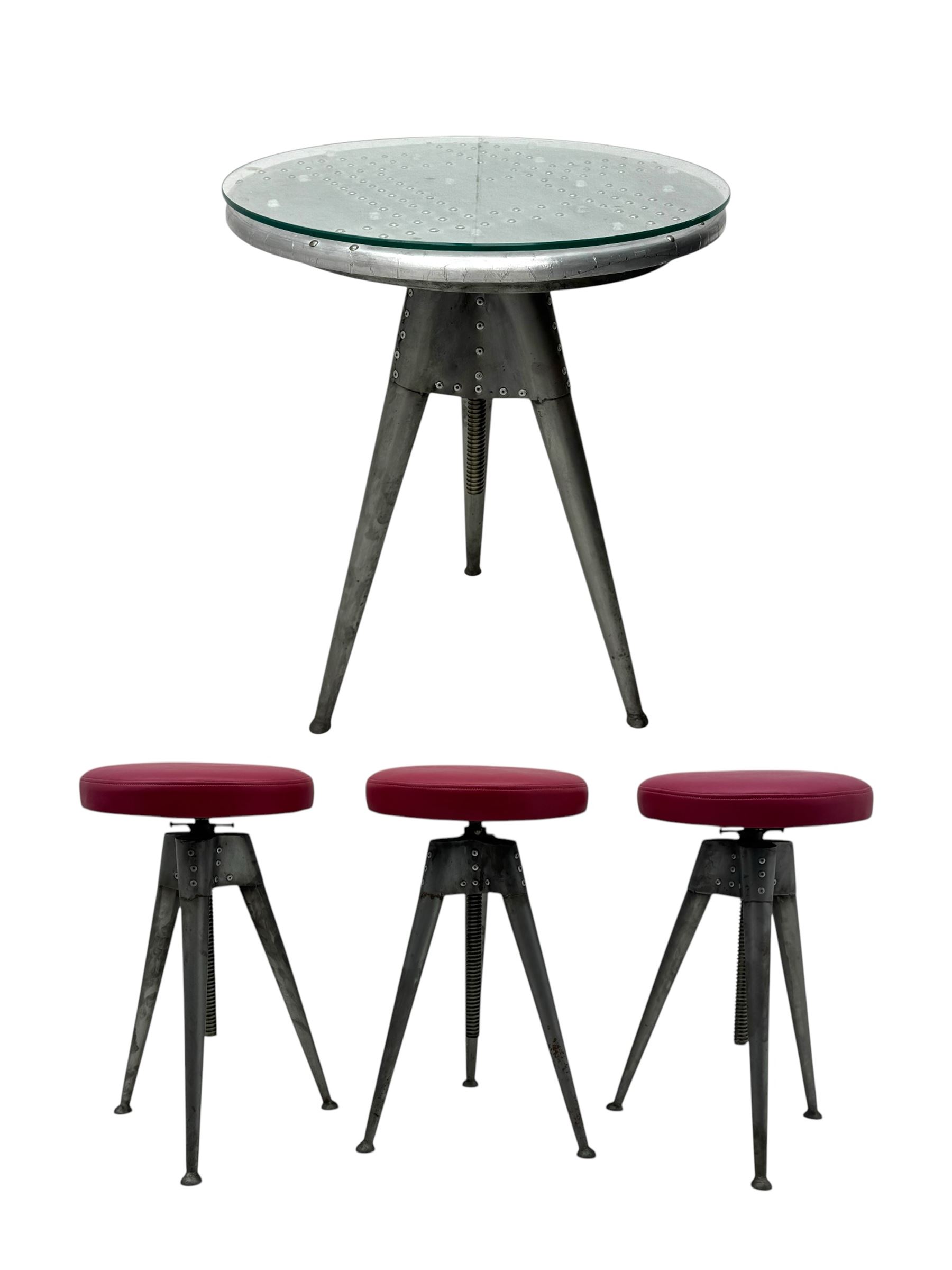 Contemporary industrial design riveted metal table, by repute fashioned from salvaged aeroplane parts, with a circular glass top on a tripod base, accompanied by three adjustable stools upholstered in burgundy