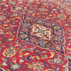 Central Persian Kashan hand-knotted rug, crimson field, profusely decorated with scrolling vines and palmettes, floral sprays around an ivory and indigo lobbed medallion enclosed by spandrels within an indigo border of arabesques between multiple guard stripes  