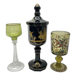 19th Century Bohemian amethyst glass lidded goblet, the bowl of faceted form, gilded with three children in period dress playing games, upon a knopped stem and conforming faceted foot, together with an art nouveau green glass goblet, with gilded rim, upon clear trumpet stem, and a hand blown wine glass, transfer printed with birds and three figures in period dress, tallest H29cm
