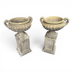 Pair of Neoclassical design weathered cast stone garden urns of kylix form, the rolled rim flanked by twin pierced scroll handles over a shallow gadrooned bowl, raised on a fluted circular socle and faux panelled square pedestal base