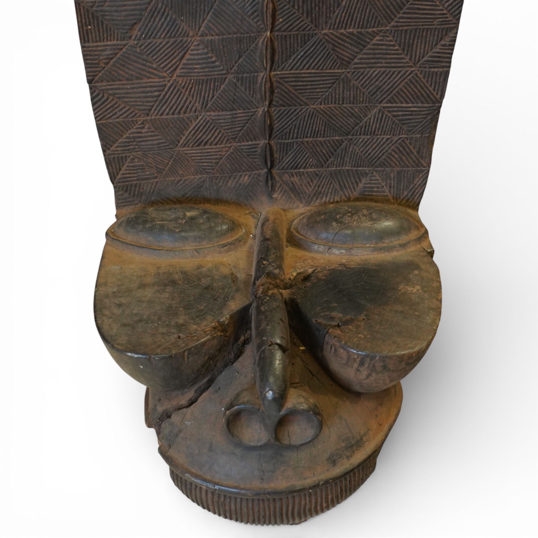African carved hardwood ceremonial seat, monoxylous construction with twin dished seat wells, the tall back incised with repeating geometric triangular motifs, the front carved in the form of a tribal mask with projecting features, raised on a cylindrical base with ribbed and textured carving
