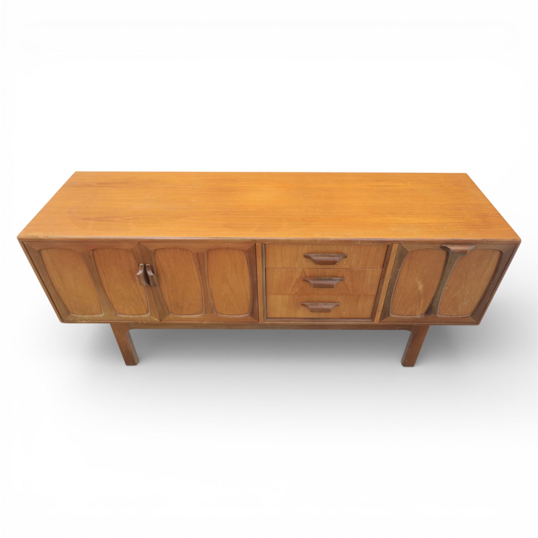 G-Plan - ‘Brasilia’ teak sideboard, rectangular top with moulded edge above a central bank of four drawers flanked by cupboard doors with carved sculptural handles, raised on tapering supports
