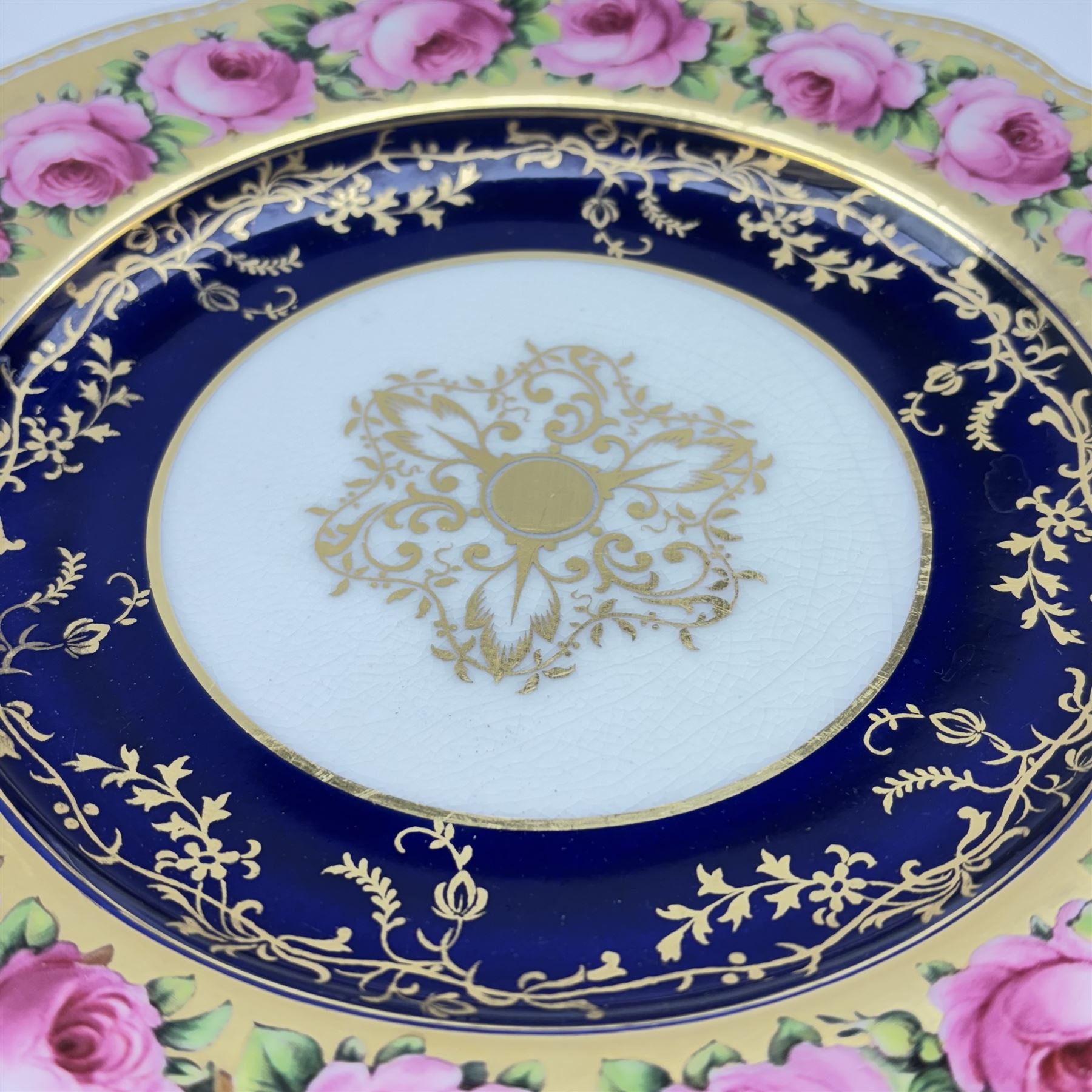 Early 19th century Coalport three handled goblet vase, shape no 114, and pair of plates, each hand painted with flowering pink roses upon a gilt band, against a dark blue ground heightened with trailing gilt tendrils, each marks beneath Coalport Swansea Period 1818, including handles vase H22.5cm, plates D23cm