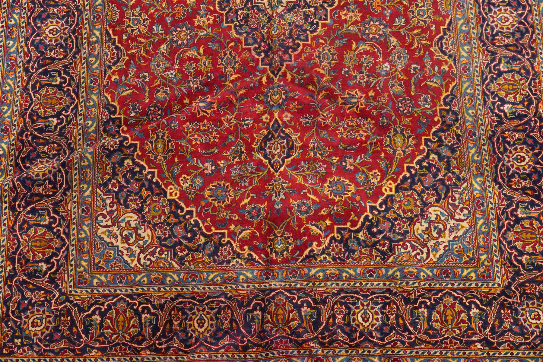 Persian Kashan hand-knotted carpet, madder red ground centred by shaped dark navy, ivory and pale blue floral medallion with pendants, surrounded by scrolling vines, palmettes and flowerheads, dark navy spandrels, enclosed by dark navy floral border with multiple floral guard stripes