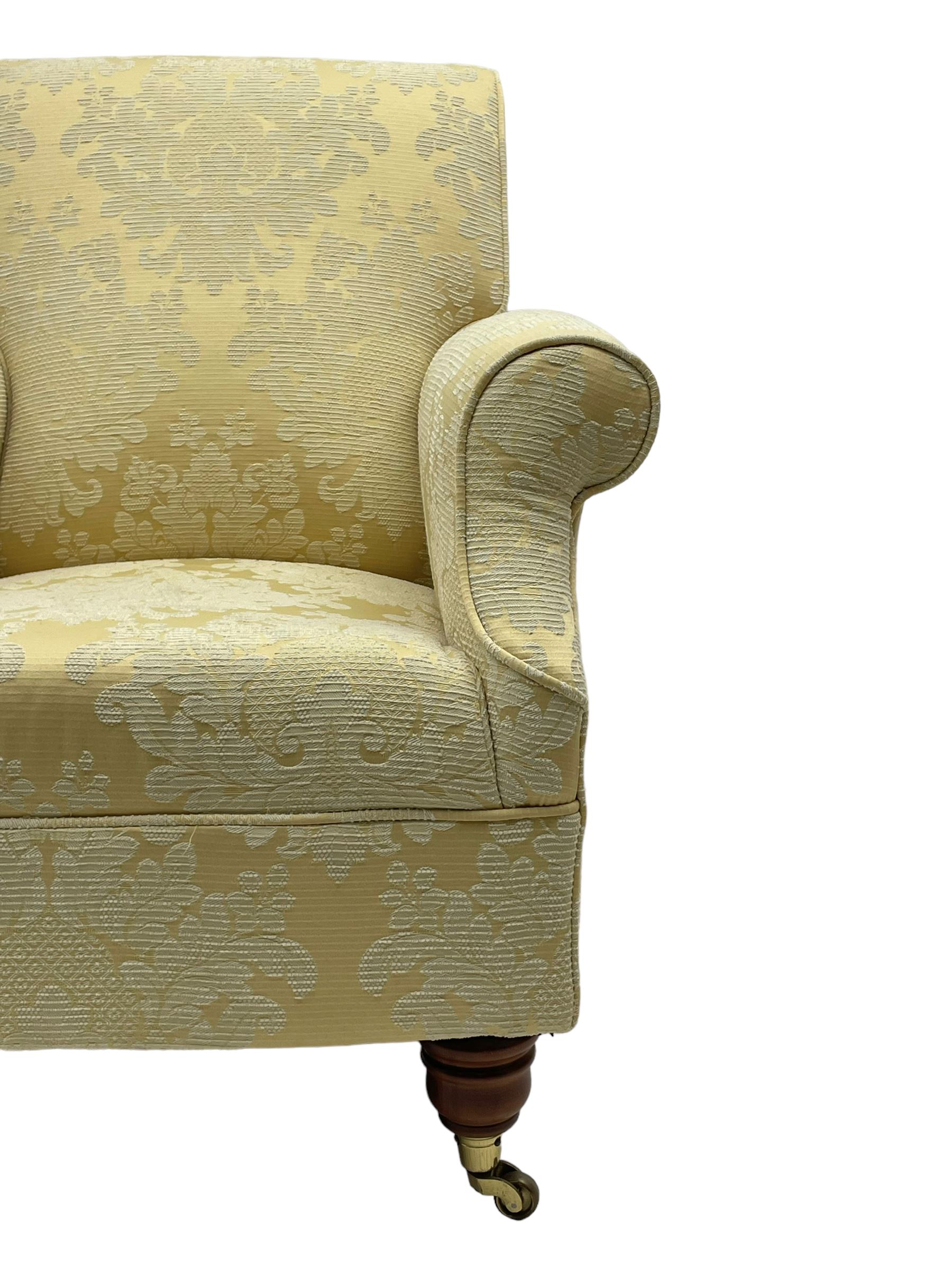 Traditionally shaped hardwood-framed armchair, rolled back and arms, upholstered in pale gold and cream damask fabric with repeating foliate pattern, on turned feet with brass cups and castors