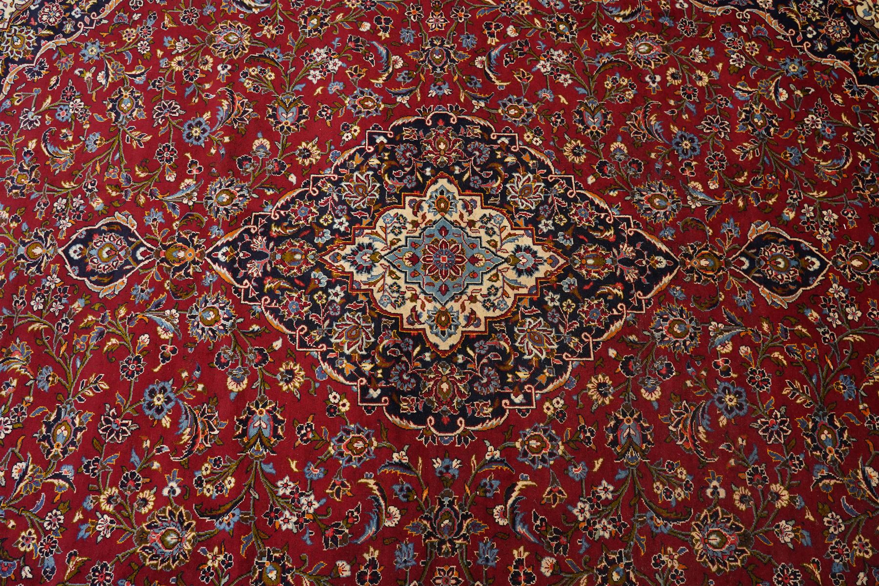 Central Persian Kashan hand-knotted carpet, crimson ground field, with central cusped lozenge medallion, enclosing a palmette and a floral rosette arrangement, surrounding field densely populated with scrolling arabesques, flowering vinery and stylized boteh motifs in tones of indigo, pale blue, ivory and ochre and spandrels of confirming design, flanked by multiple guard stripes, with cotton fringe ends 
