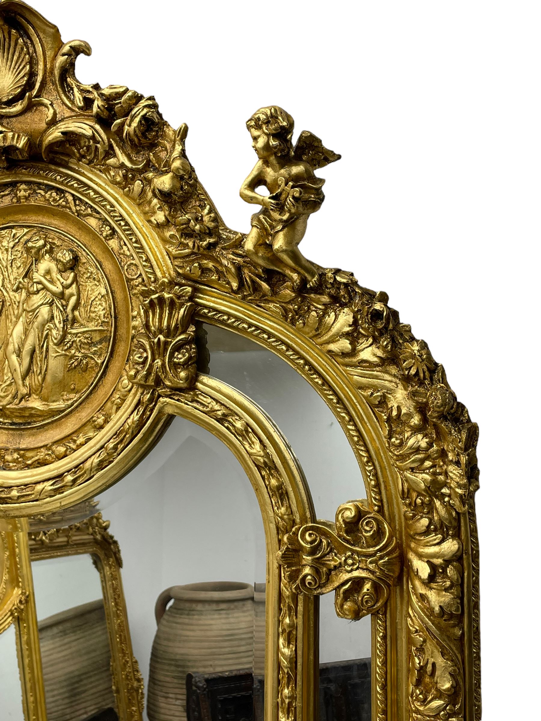 Large Italian Baroque design gilt wall mirror, the shell pediment over circular panel depicting classical female flute player with dancing putto within a tree landscape, the stepped arched upper frame surmounted by musical instrument playing putto and trailing flower heads, central bevelled mirror plate surrounded by plain segmented mirror plates, acanthus leaf moulded outer frame and foliate moulded inner slip