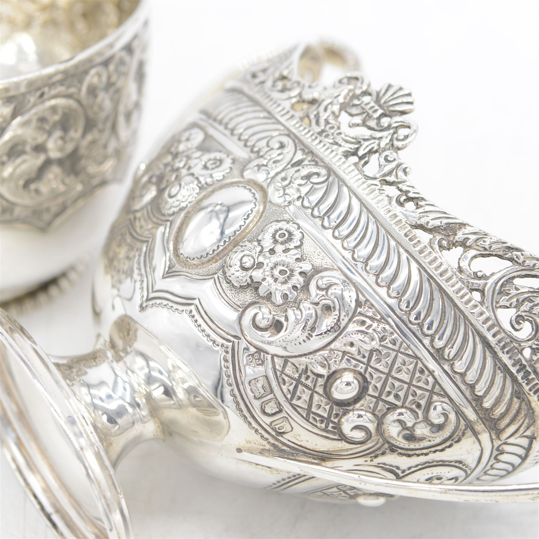 Late Victorian silver twin handled sugar bowl, profusely embossed with floral and scrolling decoration and acanthus capped handles, upon stepped oval base, hallmarked Josiah Williams & Co, London 1897, together with a Victorian twin handled sugar bowl, of circular form with embossed and chased foliate decoration and seated figures capping each handle, upon circular base, hallmarked London 1888, maker's mark indistinct, tallest H11cm