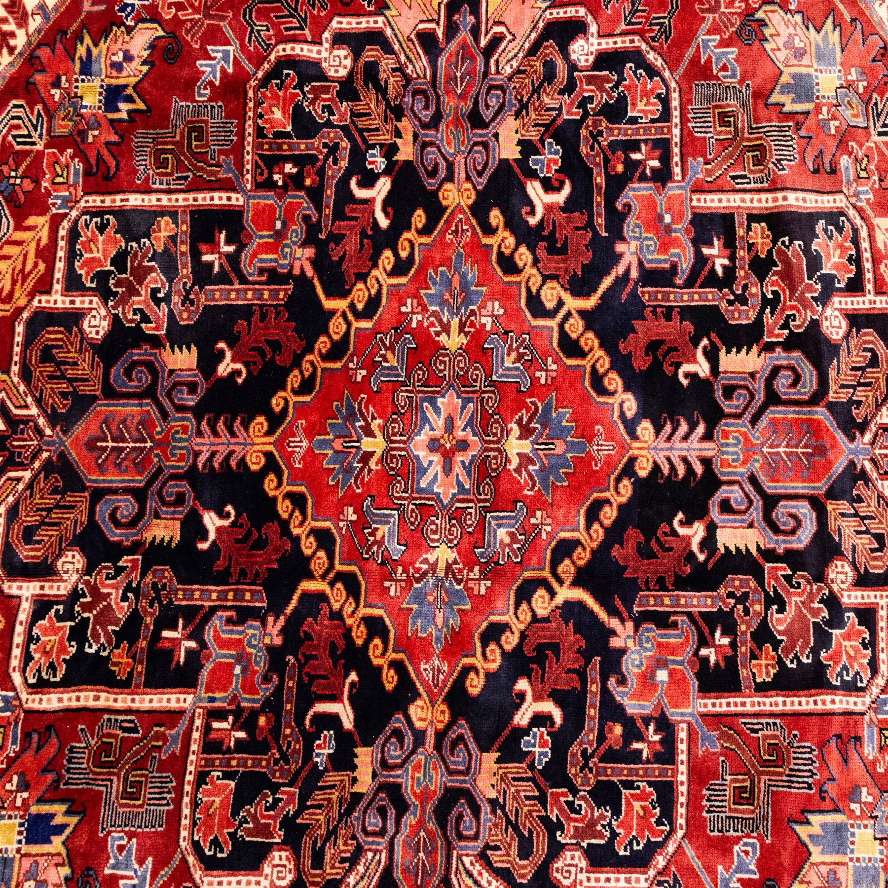 North West Persian Heriz hand-knotted carpet, madder red field with large central stepped medallion with angular floral and geometric designs, surrounded by stylized palmettes, leaves and tribal motifs with indigo corner spandrels, enclosed by a broad navy border of geometric floral devices between multiple guard stripes, with cotton fringe ends 