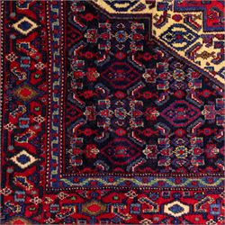 North West Persian Senneh hand-knotted rug, dark indigo field with a central bold stepped lozenge medallion of madder red and deep blue, enclosed within an ivory serrated contour forming a cruciform outline, medallion with repeating boteh and floral devices, field decorated with an all-over Herati pattern of rosettes enclosed by angular leaves principal border of madder red ground decorated with alternating floral heads, flanked by narrow guard stripes, with cotton fringe ends 