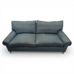 Late 20th century three seat sofa upholstered in green fabric, with gently rounded scroll arms, loose seat and back cushions, raised on turned wooden legs with castors, rear splayed legs