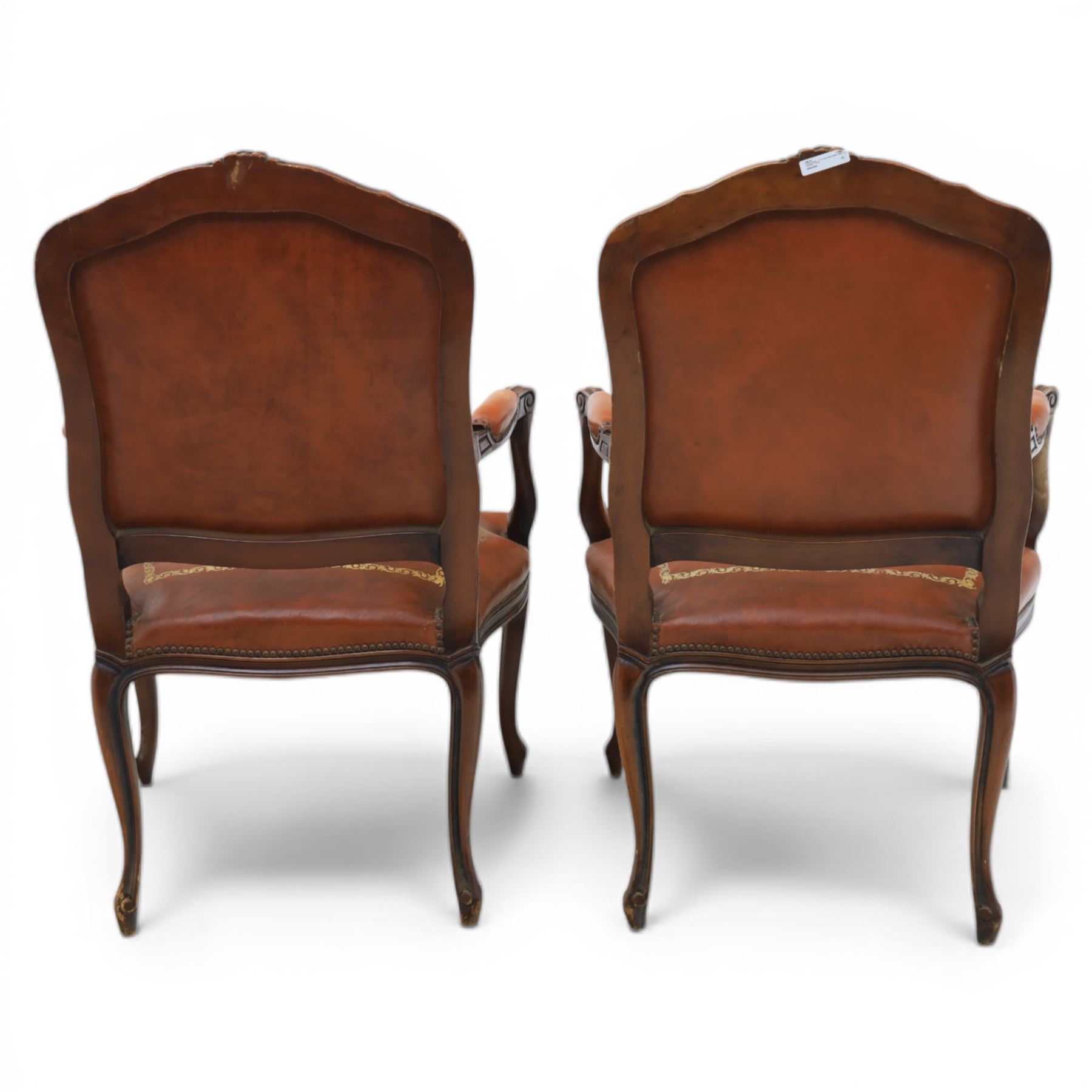 Pair of late 20th century French style carved beech open armchairs, the shaped and moulded frames with foliate cresting rail above padded backs and overstuffed seats upholstered in tan leather with gilt foliate borders and close nailed trim, raised upon cabriole supports with acanthus carved knees
