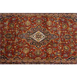 Central Persian Kashan hand-knotted rug, deep red field with central cusped ivory medallion with an all-over arrangement of scrolling floral vines, palmettes and flowering sprays within conforming spandrels, principal navy border with a continuous meandering vine of palmettes and stylized floral motifs, enclosed by multiple narrow guard borders, with cotton fringe ends 