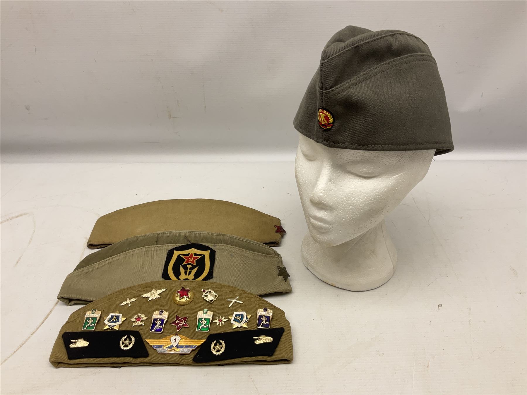 Four side caps - WW2 Russian, post-WW2 Russian and two East German; all with badges, one with multiple metal badges (4)