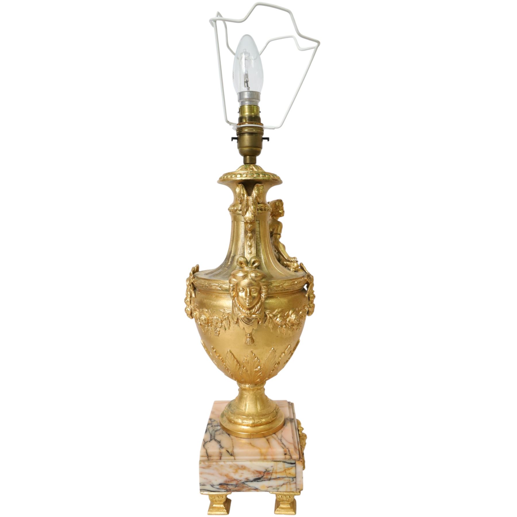Pair of Rococo style gilt metal table lamps, the urn shaped bodies each with applied figure seated upon shoulder, and female mask to opposite side issuing scroll leading to top of neck, the body cast and applied with stiff leaf detail, and foliate swags and wreaths, upon square pink marble plinths with applied gilt metal mask mounts, and four gilt metal bracket feet, each supported a pleated cream silk closed top lampshade, H63cm