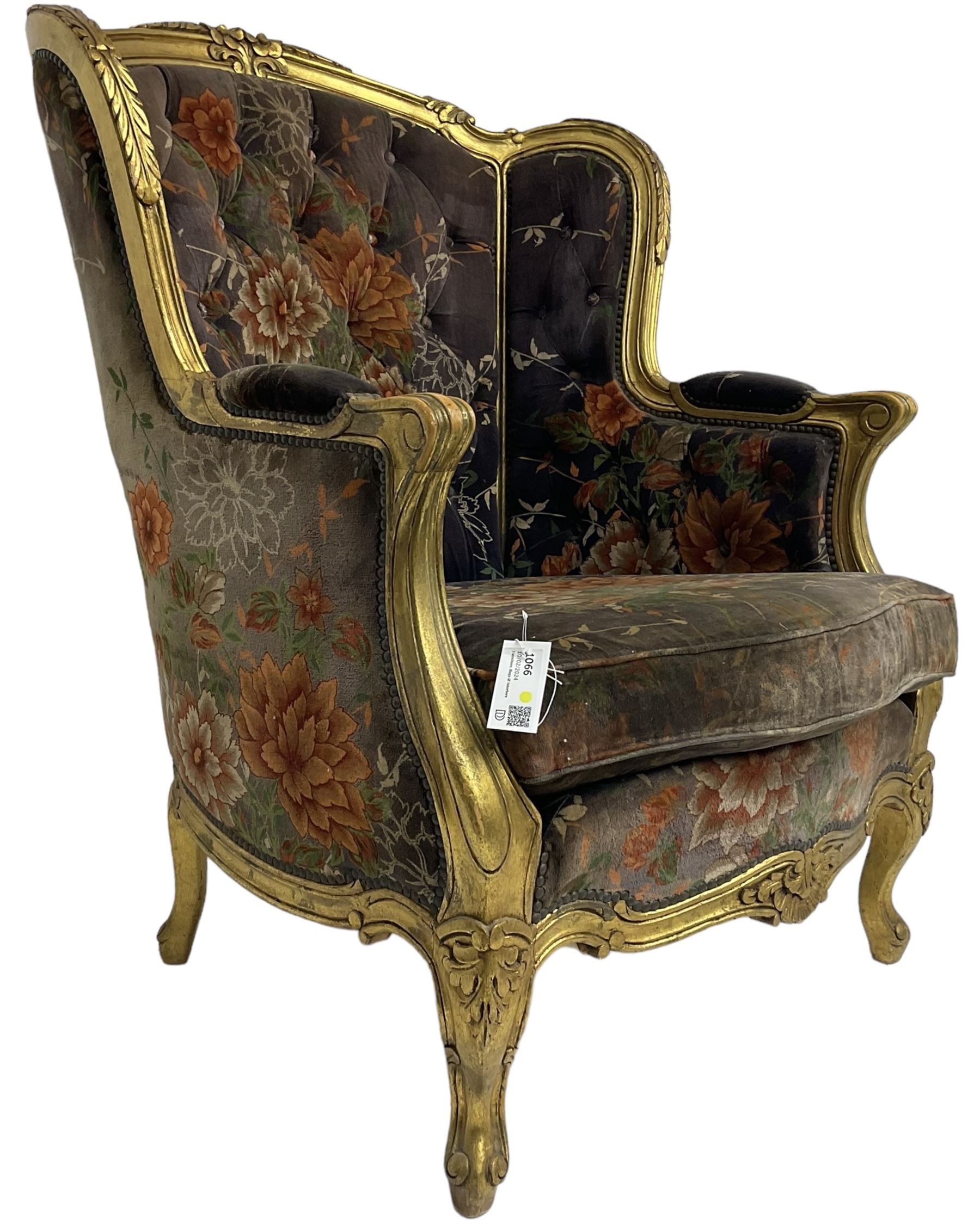 Pair of Louis XVI design gilt framed wingback armchairs, cresting rail carved and moulded with foliate decoration and C-scrolls, upholstered in buttoned floral patterned mauve velvet with loose seat cushion, scallop carved apron over cabriole supports