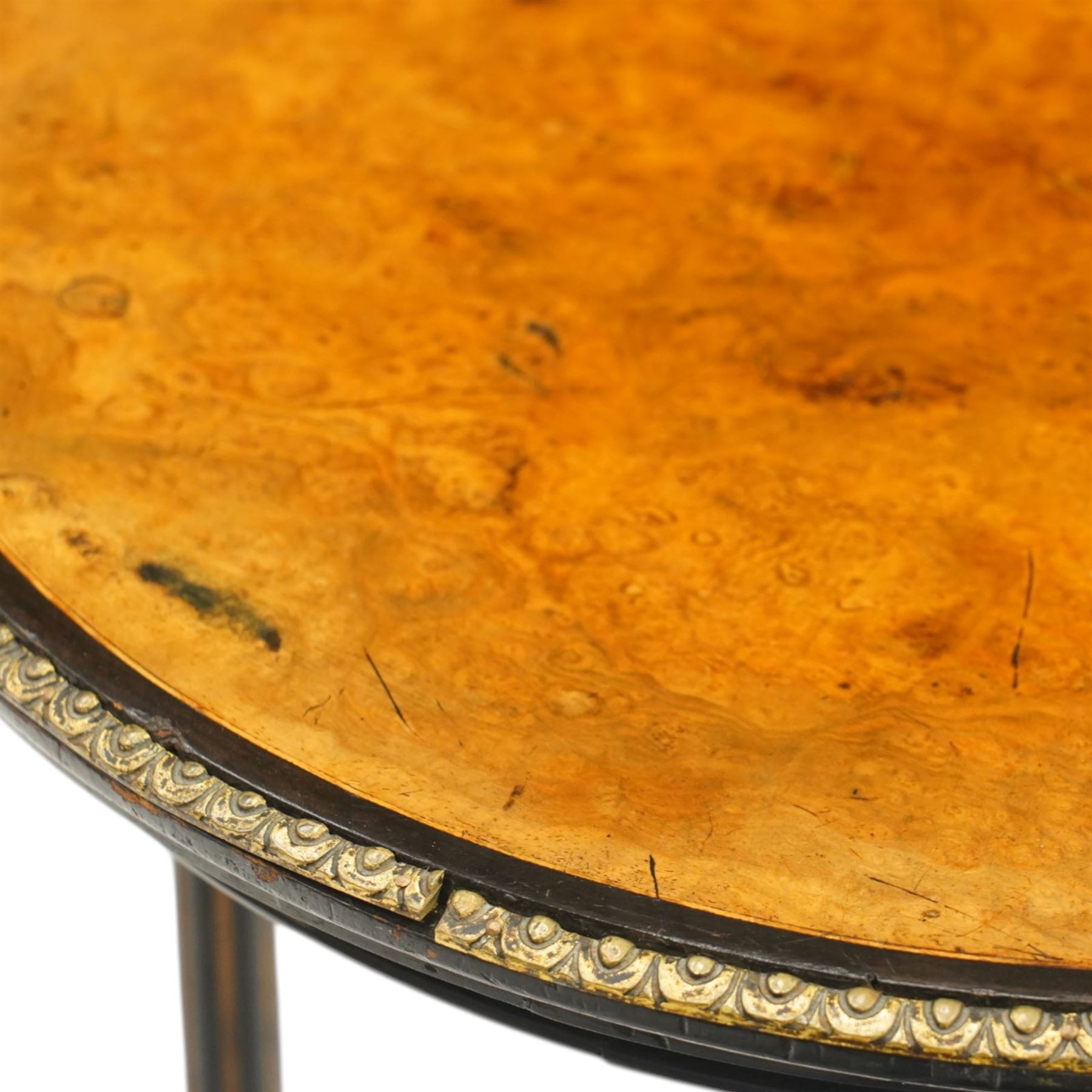 Late Victorian Aesthetic Movement ebonised and figured walnut occasional table, the circular burr top with boxwood strung and ebonised edge, surrounded by gilt metal egg-and-dart moulding, raised on three turned and fluted pedestals with parcel-gilt decoration, united by a triform undertier raised on splayed feet