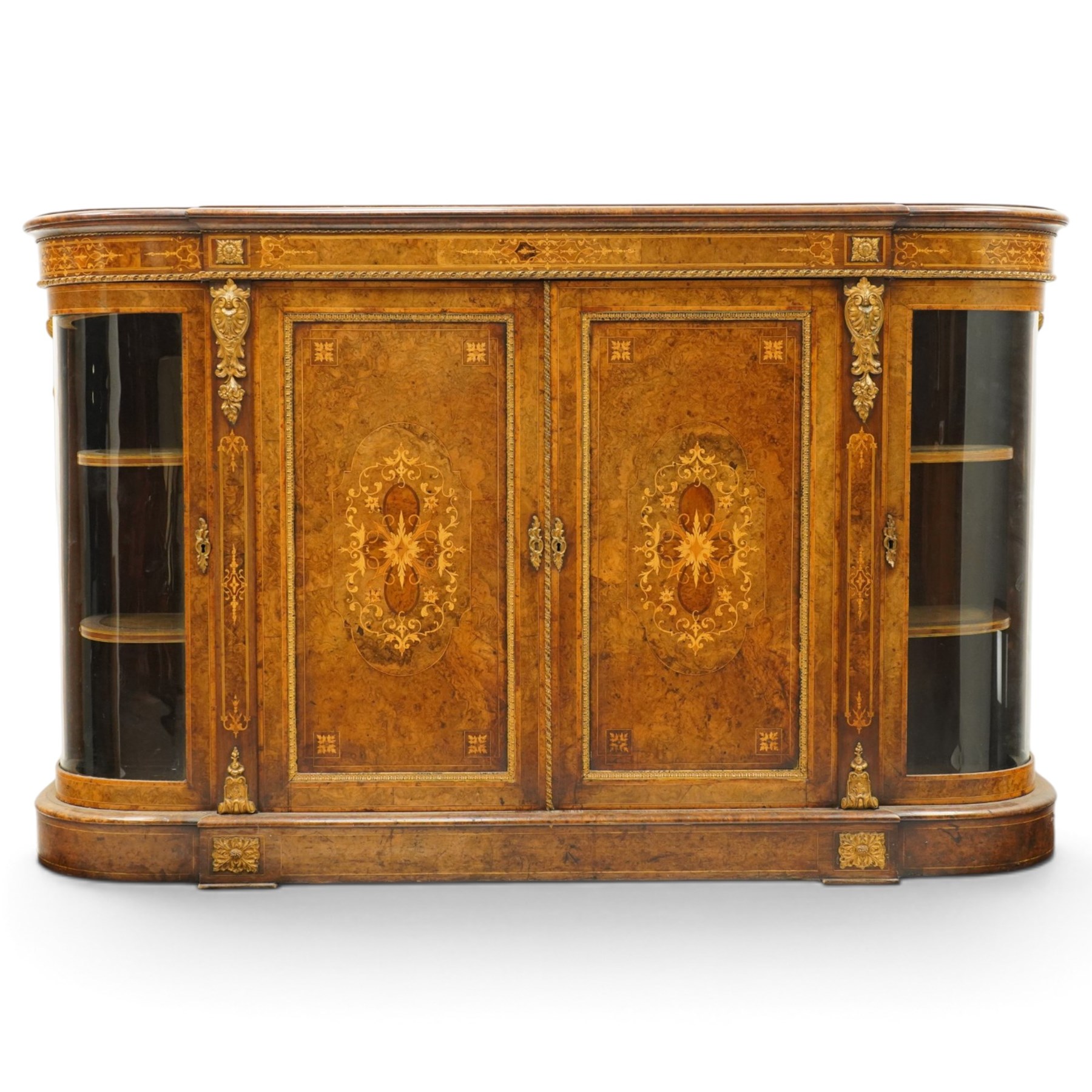 Large fine Victorian figured and burr walnut credenza, the breakfront D-shaped top with moulded edge, over an ornate frieze with inlaid Aesthetic Movement scrolling and geometric ornament, applied with gilt-metal ribbon-twist moulding and mounts, the central panelled doors profusely inlaid with scrolling foliate motifs and stringing, flanked by gilt-metal cartouche and scrolling acanthus corbel mounts, with flanking glazed bow-front side doors, on skirted base with further gilt metal foliate mounts