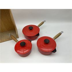 Le Creuset 'Volcanic Orange' cast iron and enamel milk pan with wood handle together with a three piece red cast iron Cousances saucepan set of graduating form, with lids housed in original wooden hanging stand and further Cousances lidded casserole dish and pan