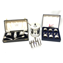  A cased silver plated set of six napkin rings and menu holders modelled as swans, together with a cased silver plated cruet set comprising pepper, open salt and mustard with blue glass liners, and spoons, a small Elkington silver plated toast rack, and a silver plated preserve jar with stylised figural pierced decoration and blue glass liner.   